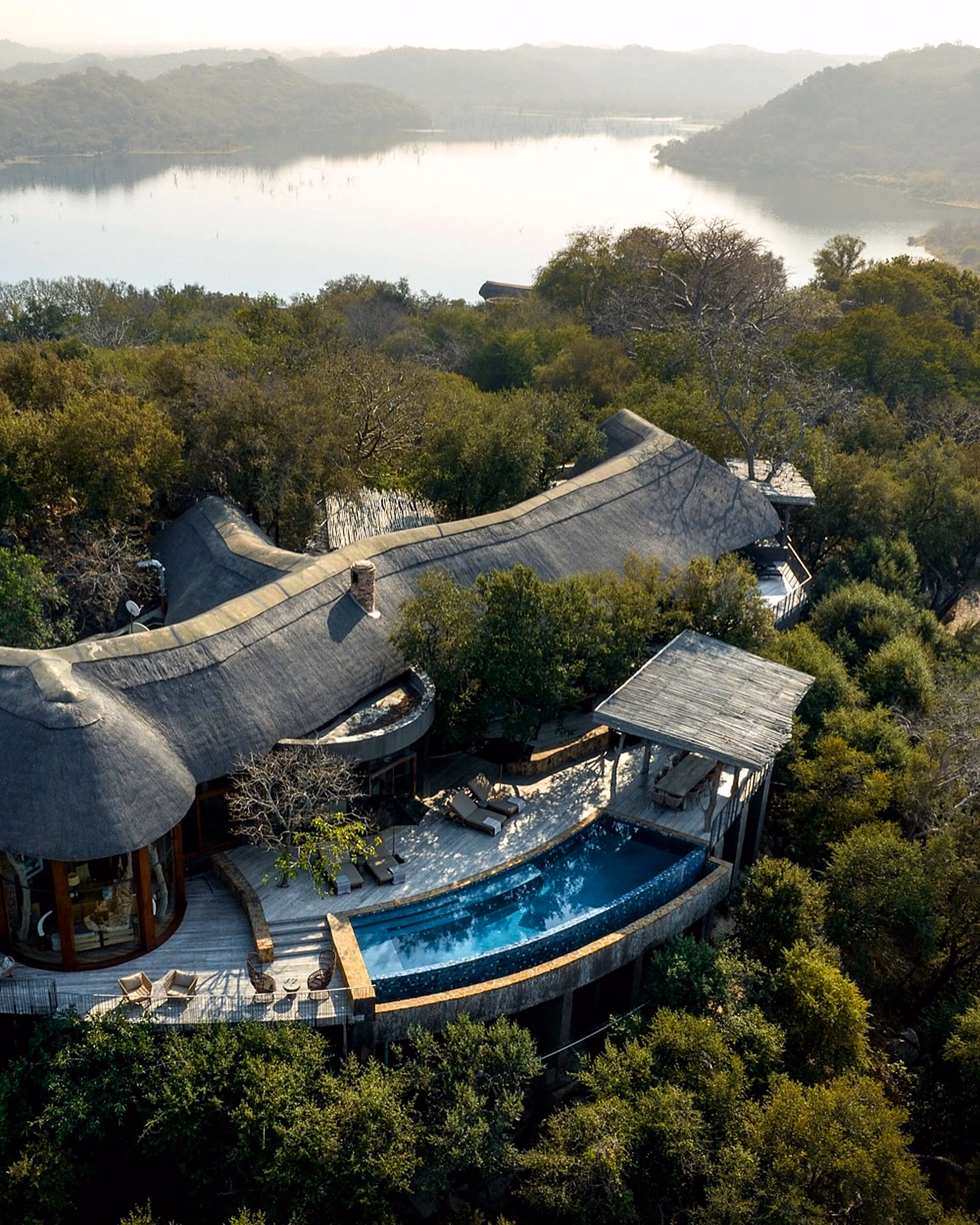 An aerial view of a curved-roof lodge and pool beside a lake.