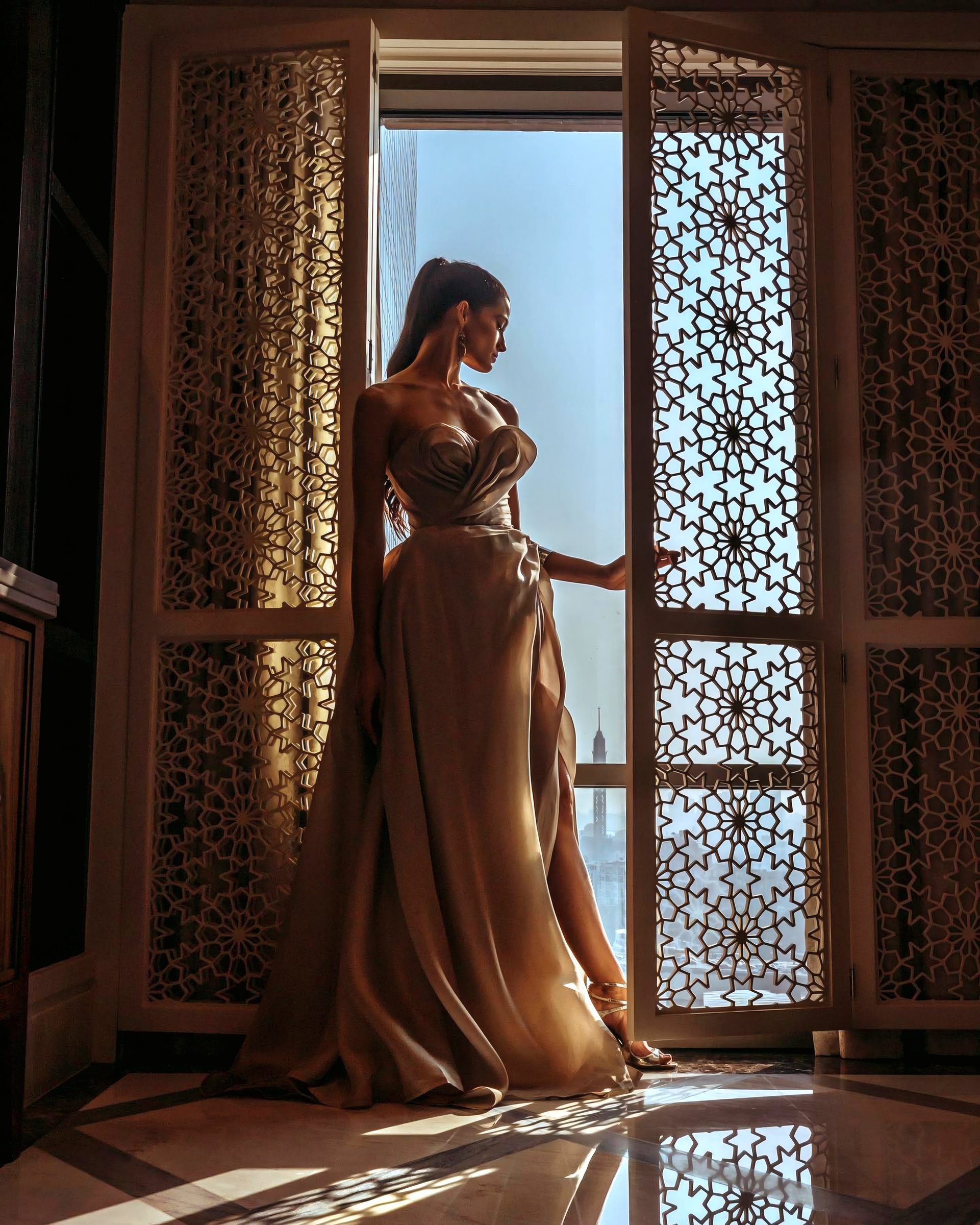 A woman standing beside an ornate sunlit window.