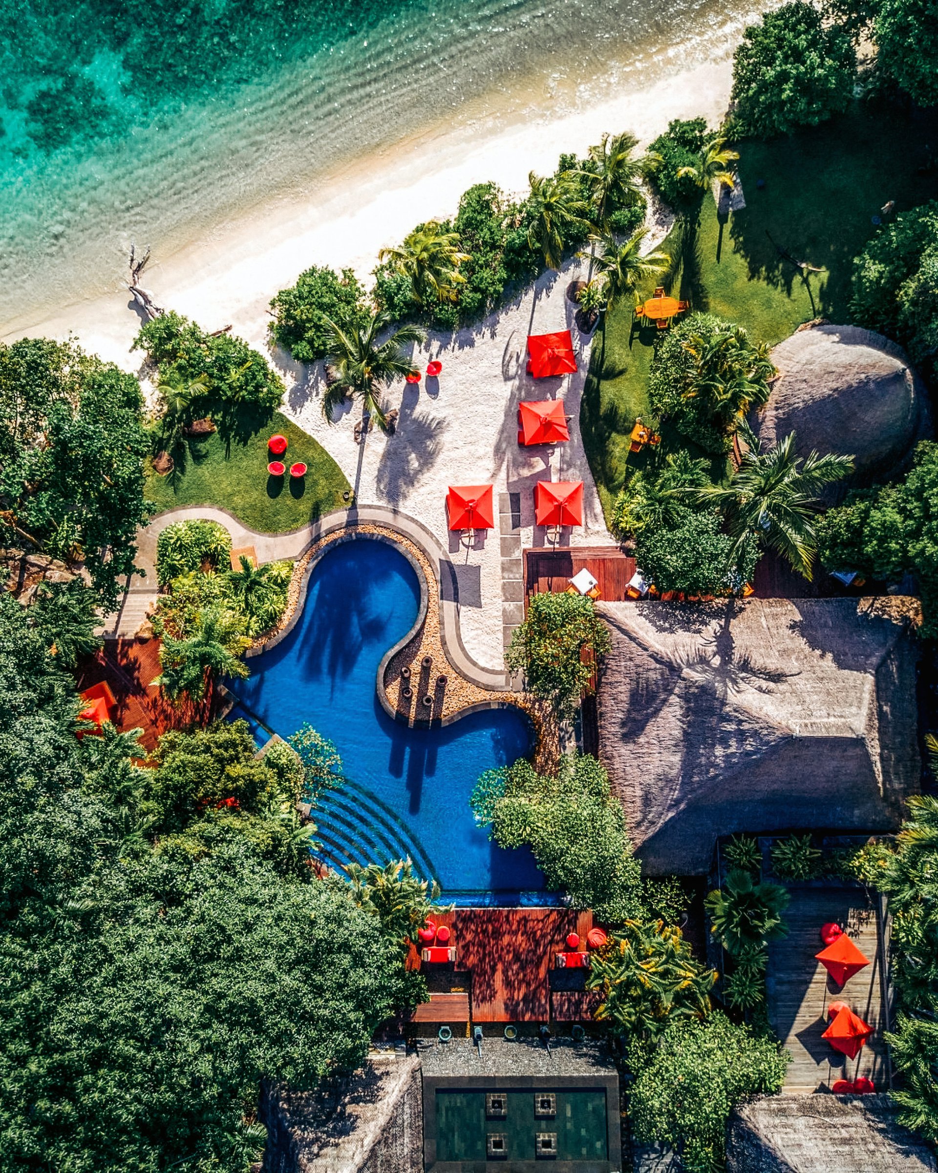 Aerial view of a tropical pool beside the beach, with red umbrellas.